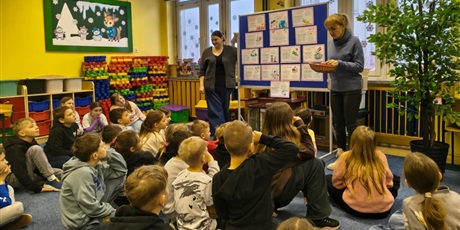 Powiększ grafikę: Grupa dzieci siedzi na dywanie w świetlicy. Wszyscy zwróceni są w stronę tablicy informacyjnej z prezentacją o walentynkach. Obok tablicy stoją dwie nauczycielki.