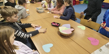 Powiększ grafikę: Grupa dzieci siedzi przy stole i wykonuje kolorowe walentynki w kształcie serc.