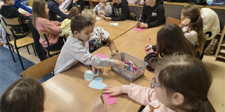 Powiększ grafikę: Grupa dzieci siedzi przy stole i wykonuje kolorowe walentynki w kształcie serc.