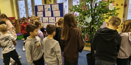 Powiększ grafikę: Grupa dzieci stoi na dywanie w świetlicy. Część z nich zawiesza serca walentynkowe na drzewie. W tle stoi granatowa tablica z prezentacją o walentynkach.