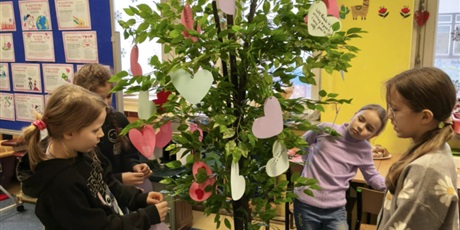 Powiększ grafikę: Grupa dzieci stoi na dywanie w świetlicy. Część z nich zawiesza serca walentynkowe na drzewie.  W tle stoi granatowa tablica z prezentacją o walentynkach.