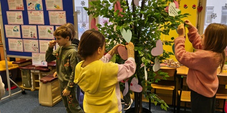 Powiększ grafikę: Grupa dzieci stoi na dywanie w świetlicy. Dwie dziewczynki zawieszają serca walentynkowe na drzewie.  W tle stoi granatowa tablica z prezentacją o walentynkach.