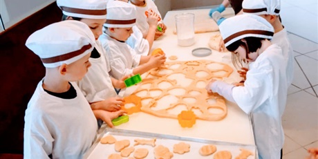 Powiększ grafikę: Dzieci wycinają ciasteczka. Dzieci noszą na sobie białe fartuchy i czepki.