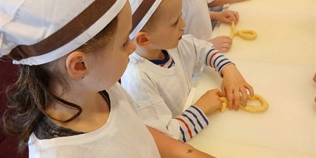 Powiększ grafikę: Dzieci w białych czepkach na głowie oraz białych koszulkach formują z ciasta precle.