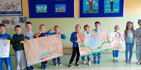 Powiększ grafikę: Uśmiechnięte dzieci stoją i siedzą przed tablicami, na których powieszone są ich prace pokazujące drzewa i kwiaty.