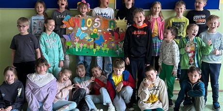 Powiększ grafikę: 6.	Chłopcy i dziewczynki stoją i trzymają kolorowy plakat z kwiatami, reszta dzieci siedzi i leży na dywanie przed plakatem.