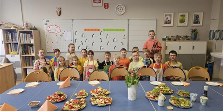 Powiększ grafikę: 7.	Na zdjęciu widzimy stół ozdobiony kwiatami w wazonie i talerze z wiosennymi, kolorowymi kanapkami. Za stołem stoją dzieci ubrane w fartuszki.