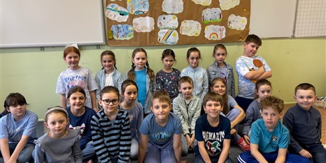 Powiększ grafikę: 13.	Uśmiechnięci uczniowie w czerwonych koszulkach stoją i siedzą przed plakatem powieszonym na ścianie, który pokazuje drzewo wokół którego fruwają motyle, a na liściach siedzą biedronki.