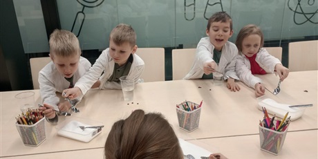 Powiększ grafikę: Dzieci ubrane w białe fartuchy wykonują doświadczenie. Łyżeczką nabierają sól i próbują ją rozpuścić w wodzie.