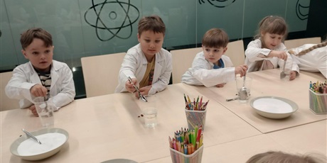 Powiększ grafikę: Dzieci ubrane w białe fartuchy wykonują doświadczenie. Łyżeczką nabierają sól i próbują ją rozpuścić w wodzie.