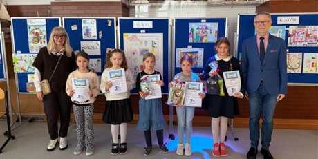 Powiększ grafikę: Laureaci konkursu "Szkoła moich marzeń" wraz z dyrekcją szkoły pozują do wspólnego zdjęcia. W rękach trzymają dyplomy i nagrody. Za nimi znajdują się granatowe tablice z pracami konkursowymi.